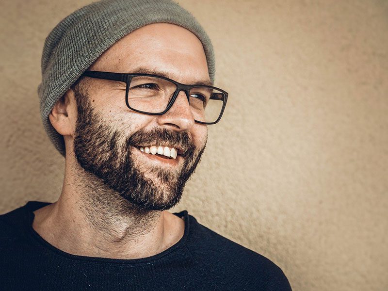 Markus Strengberger 2 Porträt eines herzlich lachenden Mannes mit Vollbart, schwarzer Brille und grauer Mütze, der zur Seite blickt