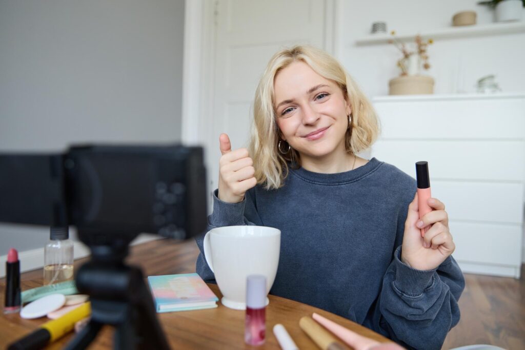 5 Content-Formate, die bei jedem Unternehmen funktionieren 2 Junge Frau (Influencerin), welche an einem Tisch vor einer Kamera sitzt, lächelt und den Daumen nach oben hält, während sie in der anderen Hand ein Makeup-Produkt in die Kamera hält.